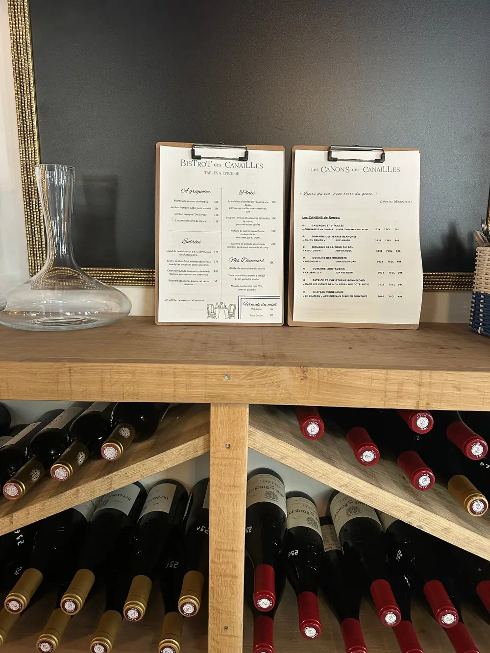 Casier à bouteilles de vin sous un comptoir en bois, avec deux cartes sur des planchettes et une carafe