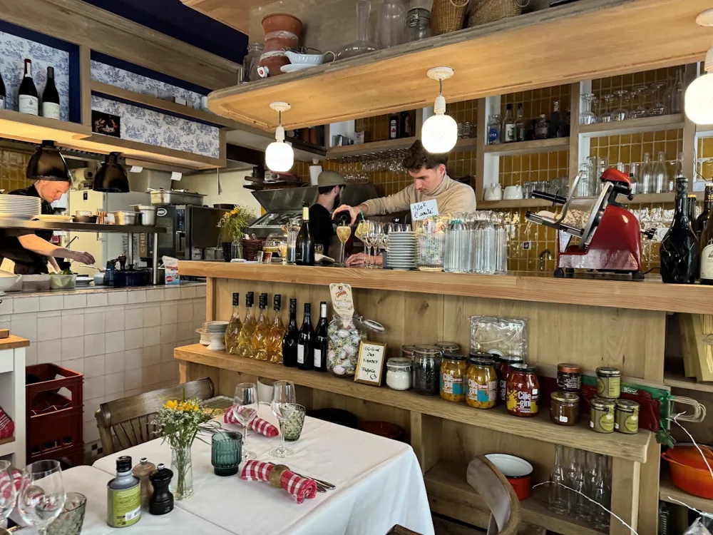 Le comptoir en bois : étagères de bocaux et de bouteilles, cuisine ouverte au fond, table dressée au premier plan