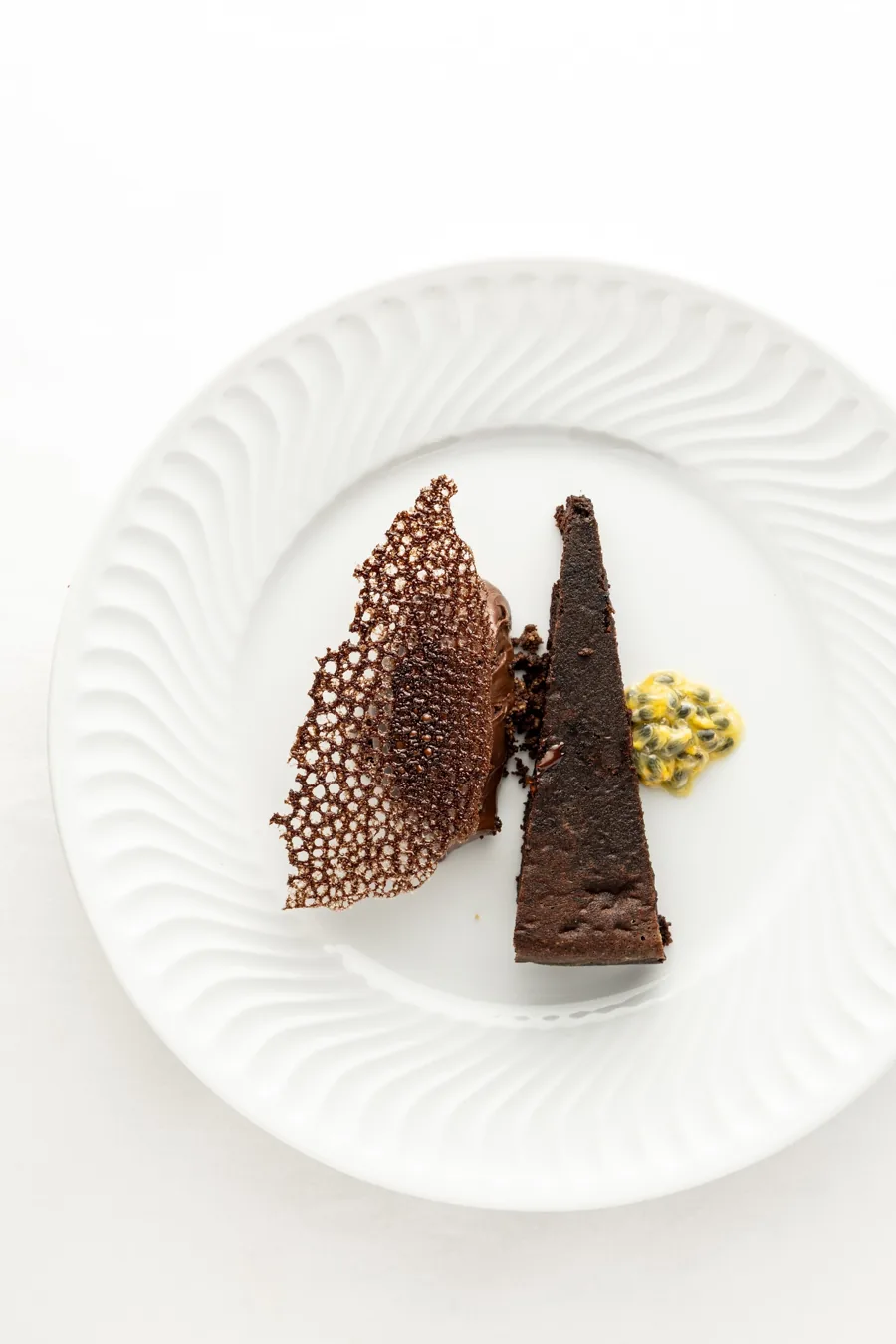 Part de gâteau au chocolat avec une tuile en dentelle et une touche de crème jaune, sur une assiette blanche cannelée
