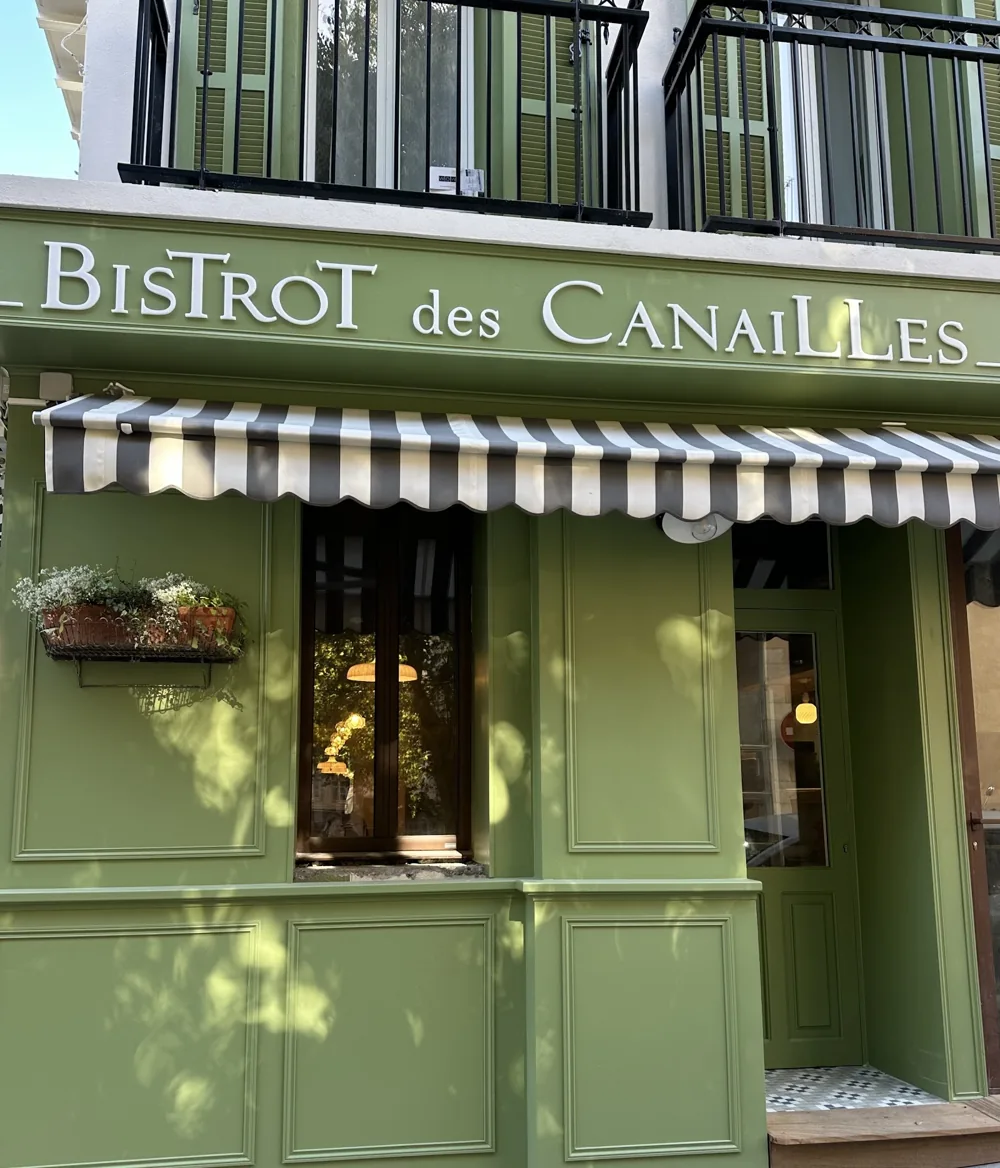 Façade verte du Bistrot des Canailles : enseigne en lettres crème et store rayé gris et blanc